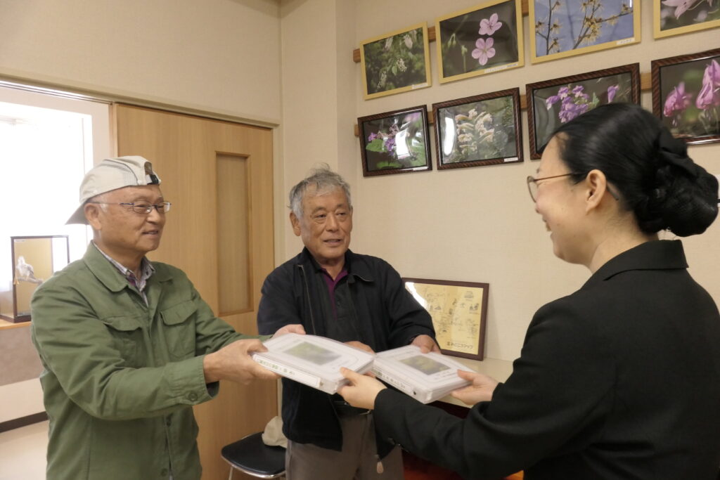 手作りの図鑑を野口館長に手渡す小林さん（左）と佐古さん