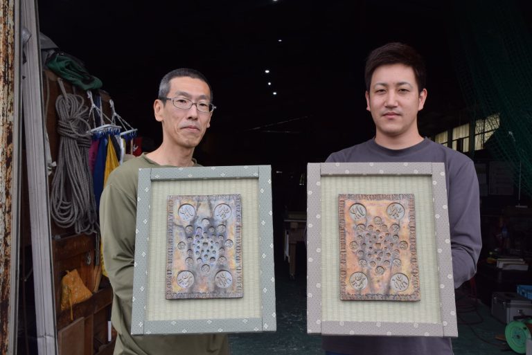 鈴木さんの新作を飾った畳表の額縁を手にする鈴木さん(左)と仲井専務