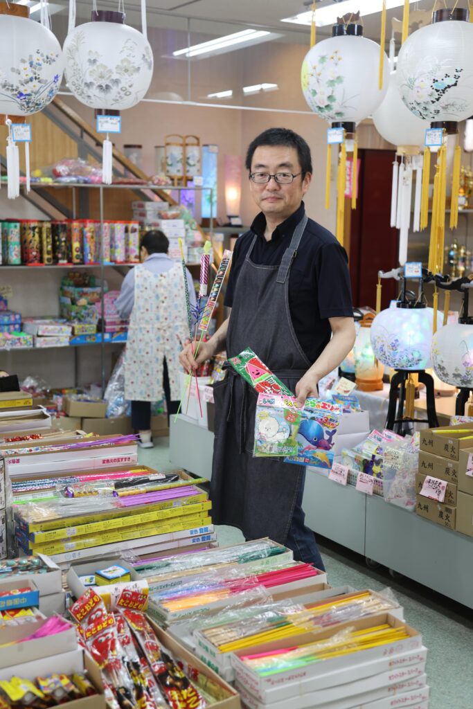 花火や盆提灯が並ぶ店内で、人気商品を紹介する店主の小坂謙一さん